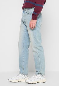Jeans de denim de lavado claro con corte recto, que presentan un diseño de cinco bolsillos, un ligero desgaste y una cinturilla estándar. Combinados con zapatillas blancas.
