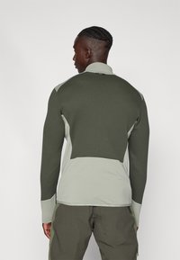 Haut de sport à manches longues bicolore en vert foncé et vert clair, avec une texture côtelée et un col montant avec un détail de couture à l'arrière.