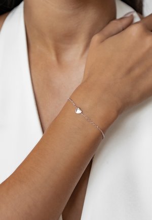 Silberne Kettenarmband mit einem herzförmigen Anhänger, glatte Textur, zartes Design, getragen an einem gebräunten Handgelenk mit einem hellen Oberteil.