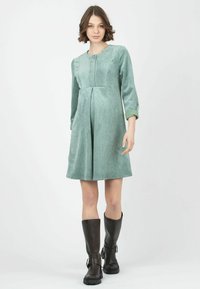 Vestido de pana verde con mangas tres cuartos y falda plisada, combinado con botas de cuero marrón hasta la rodilla.