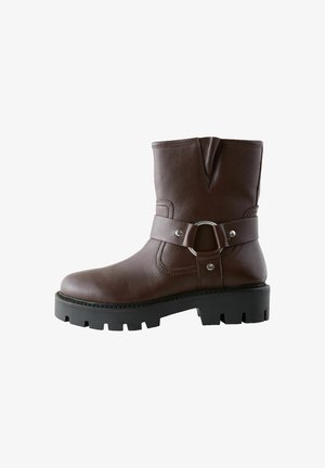 Barna bőr bokacsizma textúrázott felülettel, kerek orral, hangsúlyos csat részlettel és masszív fekete gumi talppal, amelynek barázdált mintázata van.