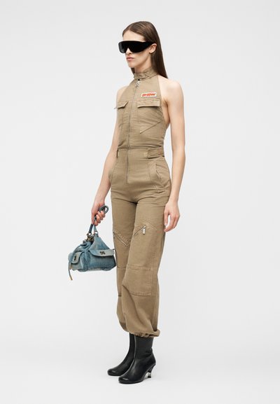 Γυναίκα που φοράει αμάνικη μπεζ ολόσωμη φόρμα με φερμουάρ στο μπροστινό μέρος, μαύρα μπότες με τακούνι, υπερμεγέθη μαύρα γυαλιά ηλίου, κρατώντας μικρή μπλε τσάντα.