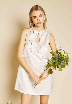 Noor naine valge varrukateta kleidis, millel on lipsud, hoiab käes roheliste lehtede ja lillade õitega kimpu beeži tausta ees.