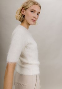 Hvit, fluffy genser med korte ermer og tettsittende liv, myk tekstur og lett gjennomsiktighet, kombinert med lyse beige skreddersydde bukser.