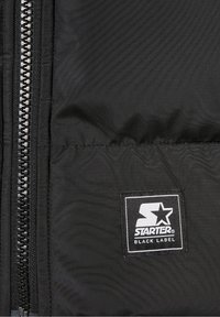 Musta puffatakki, jossa on näkyvä vetoketju ja suorakulmainen logo-tarra, jossa lukee "STARTER BLACK LABEL". Sujuva, matta kangas.