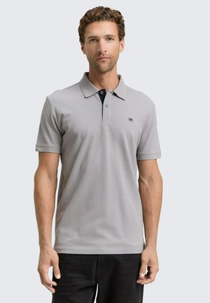Grijze polo met korte mouwen, effen textuur, knoopsluiting en klein logo op de borst. Draagt bij donkere broek.