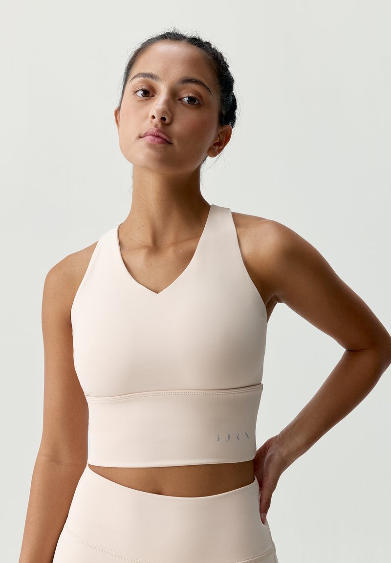 Béžový športový podprsenka s výstrihom do V, vyrobená z hladkej látky. Má široký pás a diskrétne logo na dolnom okraji.
