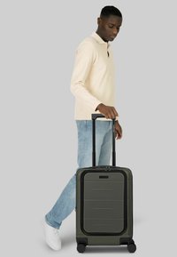 Uomo con camicia beige a maniche lunghe e jeans blu che trascina una valigia rigida verde scuro con manico telescopico e quattro ruote.