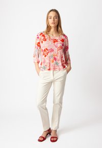 Blusa floreale nei toni del rosa, arancio e blu con maniche a 3/4; abbinata a pantaloni bianchi sartoriali e sandali con tacco rossi. Sfondo semplice.