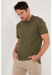 Camisa polo de canalé verde oliva con cuello, tapeta de tres botones y manga corta. Tejido texturizado con una silueta ajustada.