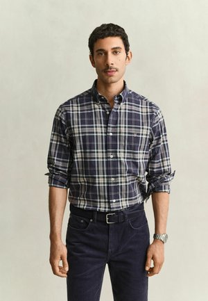 Man draagt een marineblauw-wit geruit overhemd met opgerolde mouwen, donkere broek, zwarte riem en een zilverkleurig polshorloge, staand tegen een effen achtergrond.