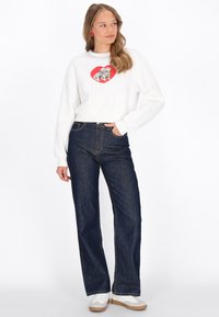 Hanorac alb cu un grafic roșu în formă de inimă, care prezintă doi câini cu pete, asortat cu blugi evazați albastru închis și adidași albi.