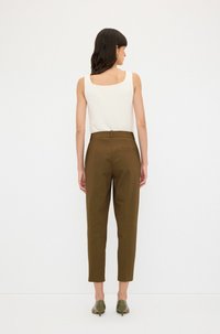 Hnědé kalhoty na míru s vysokým pasem a slim fit, doplněné bílým tílkem bez rukávů. Modelka má na sobě zelené podpatky.