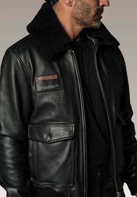 Homme portant une veste bomber en cuir noir avec col en fourrure et détails en écusson, tourné sur le côté contre un fond neutre.