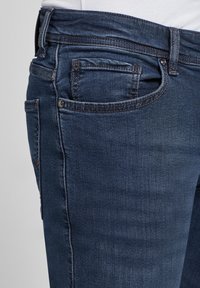 Mörkblå denimjeans med en figursydd design, som har fem fickor, kontrasterande sömmar och en klassisk metallknappstängning.