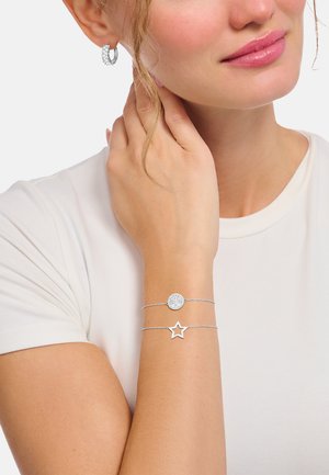 Donna che indossa braccialetti con catena a stella e ciondolo rotondo in argento e orecchini a cerchio in argento, si tocca i capelli, indossa una maglietta bianca a maniche corte.
