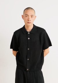 Black knitted short-sleeve shirt with a button-up front, featuring a textured pattern and a classic collar. Silhouette is relaxed-fit.
