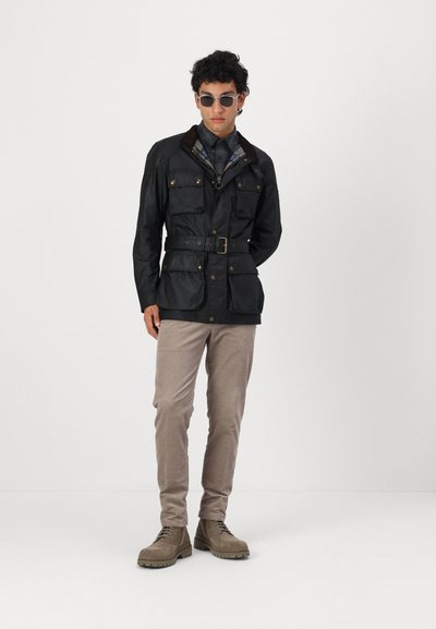Veste en coton ciré noir avec quatre poches frontales, taille ceinturée et col en velours côtelé marron, assortie à un pantalon beige clair et des bottes marron.