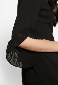 Svart klänning med detaljer i veckad, genomskinlig ärm med vågig kant, vilket framhäver en kontrast i textur och design.