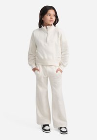 Krémová mikina na zips so voľným strihom, doplnená zodpovedajúcimi širokými nohavicami. Materiál sa zdá byť mäkký a textúrovaný, s minimalistickými dizajnovými detailmi.