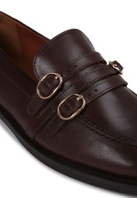 Brown leather loafer featuring two decorative gold buckles, stitched detailing along the upper, and a flat black sole. Smooth texture.