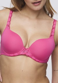 Sostén acolchado rosa con borde de encaje festoneado en las tirantes, un lazo decorativo en el centro y una textura suave y lisa en todo.