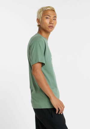 Grøn bomulds t-shirt med kortærmer, rund hals og hvid tekstgrafik på forsiden. Modellen har sorte bukser på.