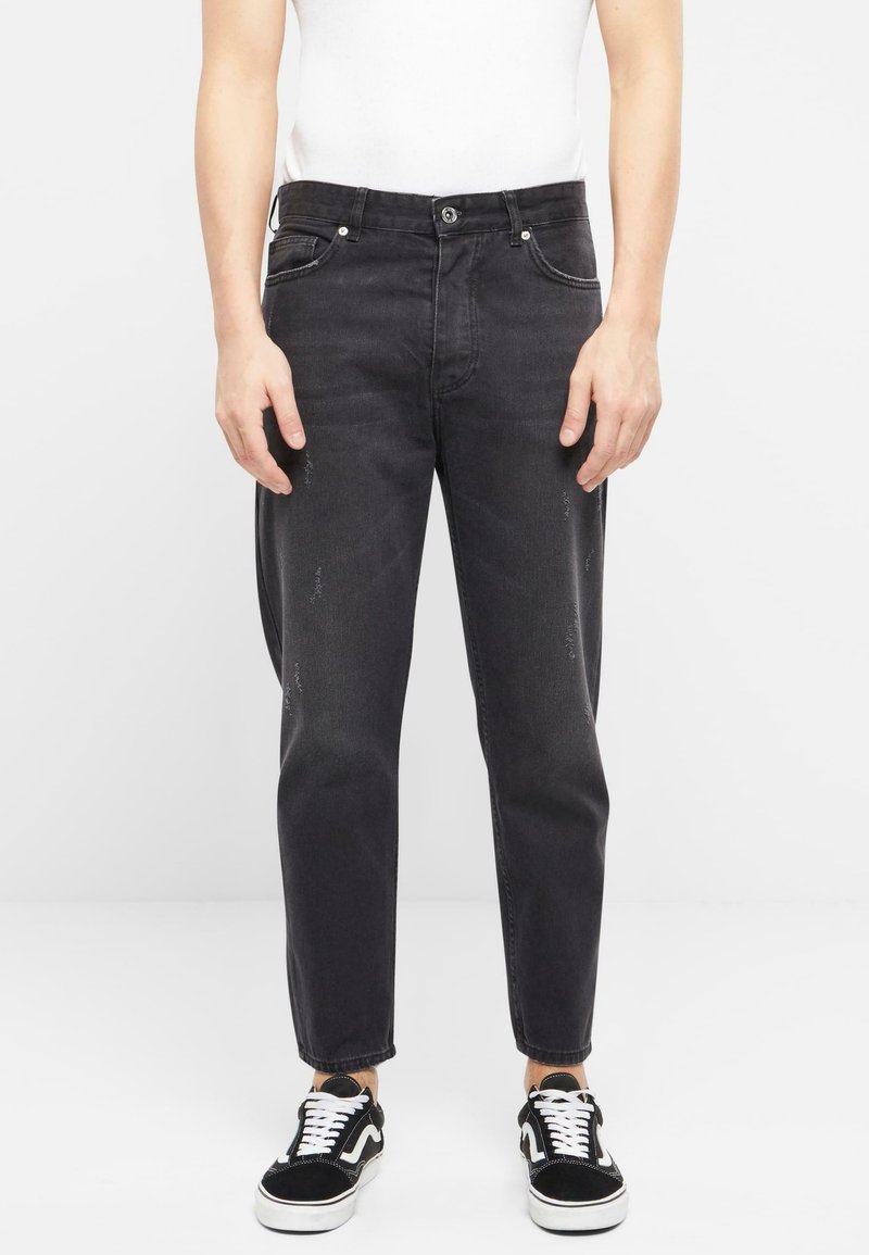 Pantalones vaqueros de mezclilla negra con corte recto, que cuentan con un cierre de botón, bolsillos frontales, detalles sutilmente desgastados y bordes deshilachados. Combinados con zapatillas negras.