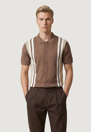 Giovane uomo che indossa una camicia marrone a maniche corte con bottoni e righe verticali bianche, e pantaloni marrone scuro, in piedi con le mani nelle tasche.