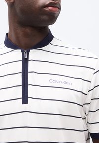 Άνδρας που φοράει λευκό και ναυτικό μπλε ριγέ φούτερ Calvin Klein με φερμουάρ, σκούρο γιακά και εμφανές λογότυπο της μάρκας στο στήθος.