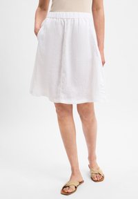 Nainen, jolla on yllään valkoinen polvipitkä hame, kädet taskuissa ja beige kierre solmuton sandaalit valkoista taustaa vasten.