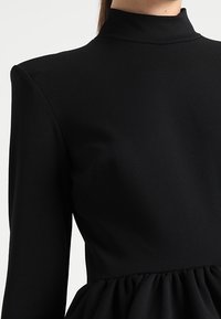 Musta mekko, jossa on korkea kaulus ja pitkät hihat. Bodice on istuva, ja hame on kerätty, luoden tekstuurisen ja valuva ilmeen.