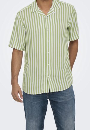 Mężczyzna w krótkim rękawie, koszuli na guziki w pionowe zielono-białe paski oraz w niebieskich dżinsach, z rękami swobodnie opuszczonymi wzdłuż ciała.