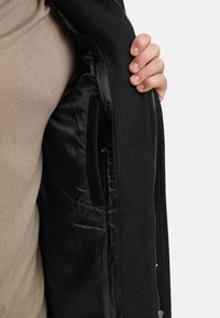 Svart glidelåsjakke med ribbet tekstur og glatt satinfôr. Synlig innerlomme. Hånd vist som holder jakken åpen.