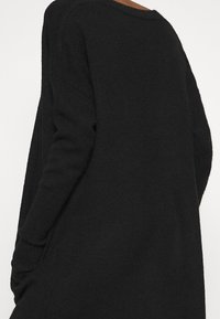 Black oversized sweater with a textured finish, boat neckline, and wide sleeves, featuring side slits and a relaxed fit design.