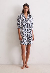 Mujer de pie descalza contra una pared blanca, vistiendo un vestido de camisa de noche de manga larga, abotonado y con estampado floral.