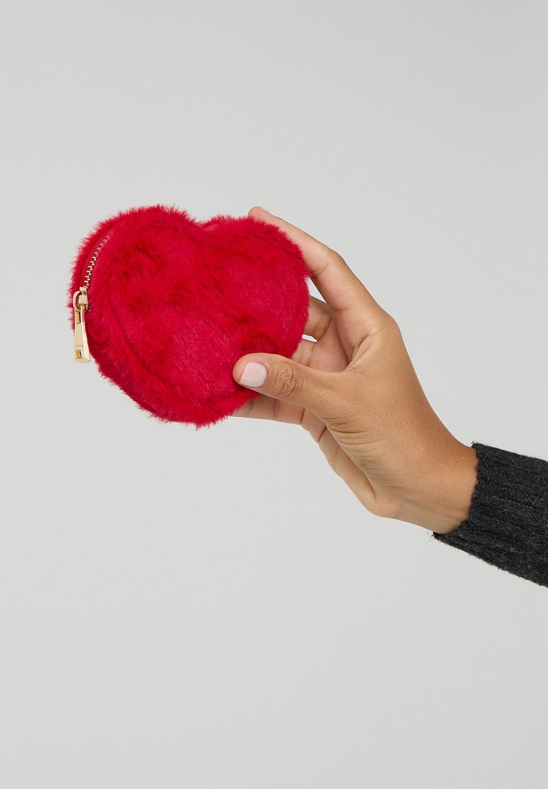 Borsa a forma di cuore rossa realizzata in morbido materiale plush; presenta una zip dorata ed è tenuta in una mano con un maglione nero strutturato.