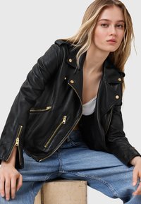 Giacca da biker in pelle nera con zip e dettagli dorati, caratterizzata da un frontale asimmetrico e spalle strutturate. Design testurizzato e aderente.