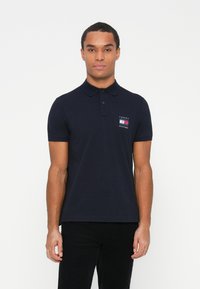 Mann iført en marineblå Tommy Hilfiger polo med logo på venstre bryst, stående mot en enkel hvit bakgrunn.