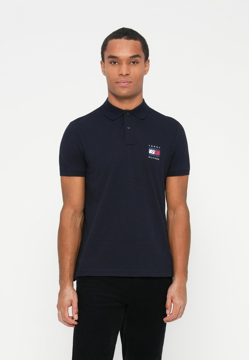 Mann iført en marineblå Tommy Hilfiger polo med logo på venstre bryst, stående mot en enkel hvit bakgrunn.