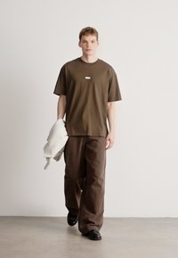 Brązowy, oversize'owy t-shirt z krótkimi rękawami i małą metką z przodu, w połączeniu z luźnymi, ciemnobrązowymi spodniami i czarnymi butami.