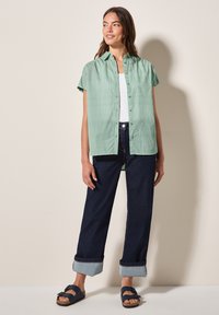 Camicia a maniche corte a righe verdi con un frontale a bottoni, indossata sopra un top bianco. Abbinata a pantaloni di denim scuri e sandali blu navy.