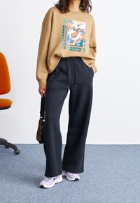 Felpa oversize beige con stampa grafica, abbinata a pantaloni della tuta neri. Scarpe da ginnastica viola chiaro e bianche, e una piccola borsa con motivo.