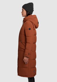 Bruine pufferjas met een capuchon, zijkleppen en een gestikt ontwerp. Gecombineerd met een donkergrijze beanie. De stof lijkt zacht en glad.