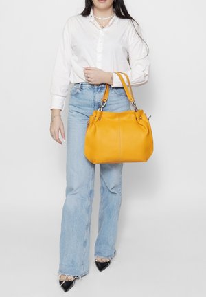 Yellow leather handbag with a rounded shape, featuring two handles and silver hardware. Smooth texture with visible stitching details, held by a person.