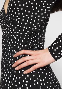 Svart klänning med vita prickar, omslagdesign med knuten midja och långa ärmar. Slät tygtextur med figursydd silhuett.