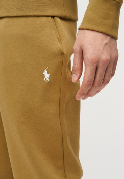 Χέρι που ακουμπά κοντά στην τσέπη σε κίτρινες φόρμες με λευκό κεντημένο λογότυπο παίκτη πόλο.