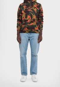Sudadera camuflada en verde oscuro, naranja y marrón; presenta un bolsillo tipo canguro y capucha con cordón. Combinada con jeans azul claro y zapatillas blancas.