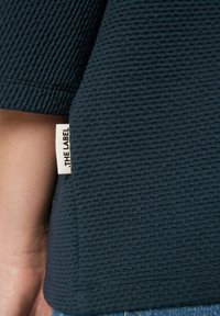 Dunkelblaues, strukturiertes Gewebe mit einem subtilen Gittermuster; zeigt ein sichtbares Etikett mit "THE LABEL" in schwarzer Schrift auf einem cremigen Hintergrund.