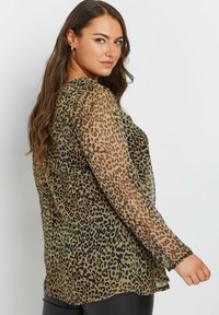 Blusa leopardata trasparente con maniche lunghe, caratterizzata da un mix di pattern marroni e neri su uno sfondo oliva. Design leggero e fluido.